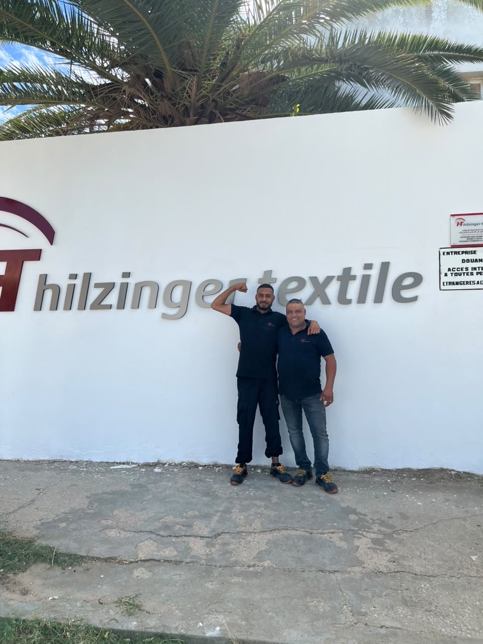 Deux collègues devant la marque Hilzinger Textile, l’un fléchissant le bras dans un geste de force